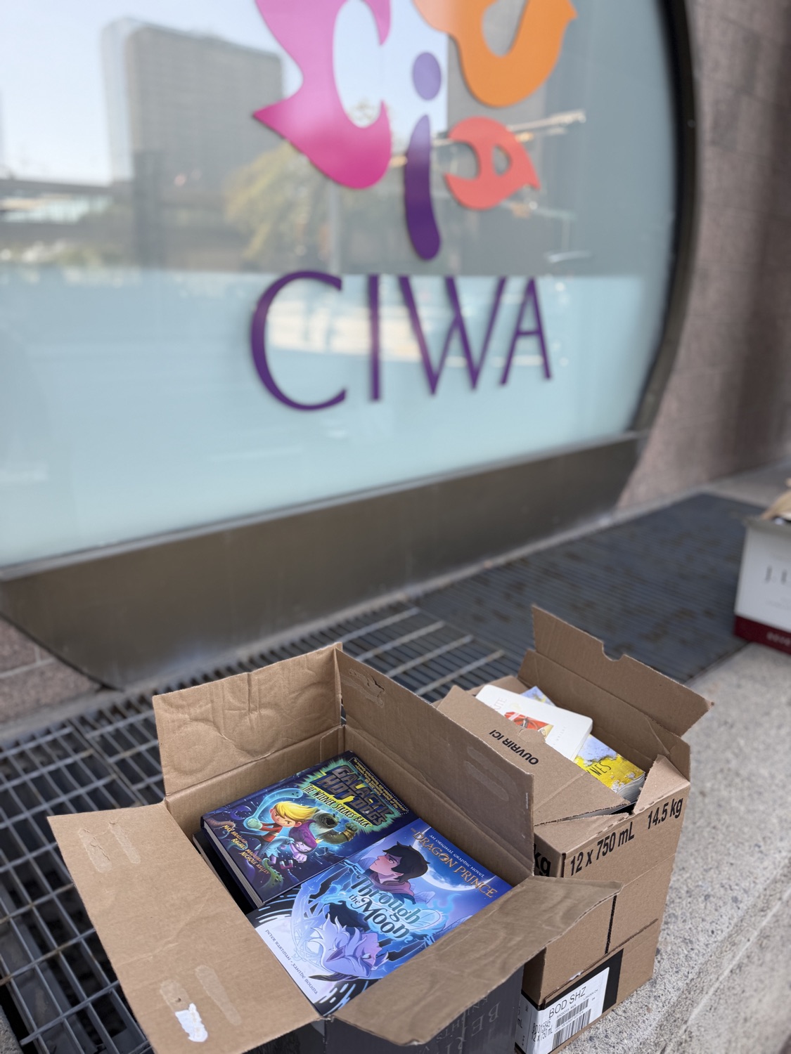 An open box of children's books at CIWA's door, with the CIWA logo in the window above